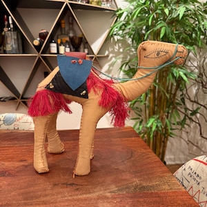 Op de afbeelding: Een bruin stoffen kameel met een blauw en zwart zadel, rode franjes en een groene touwteugel. De kameel heeft gestikte details, waaronder een oog. De kameel staat op een houten tafel.