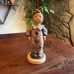 May include: A vintage ceramic figurine of a young clown in a lavender and yellow outfit with colorful buttons. The figurine has a surprised expression and is standing on a round, cream-colored base. The clown is holding a small red object.