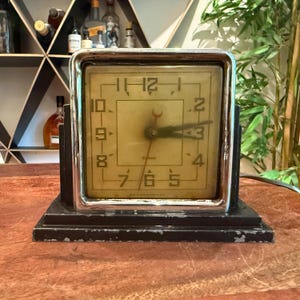 Puede incluir: Un reloj eléctrico cuadrado vintage con marco cromado y base negra. La esfera es de color blanco roto con números y manecillas doradas. La marca Gilbert está impresa en la esfera. La base muestra signos de desgaste y desconchado.