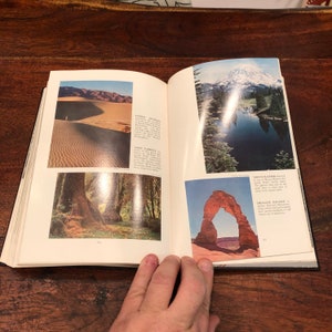 May include: An open book displaying colourful images of various landscapes. The book features pictures of sand dunes, a forest, a mountain with a lake, and a natural arch. The book's pages contain text describing the locations.