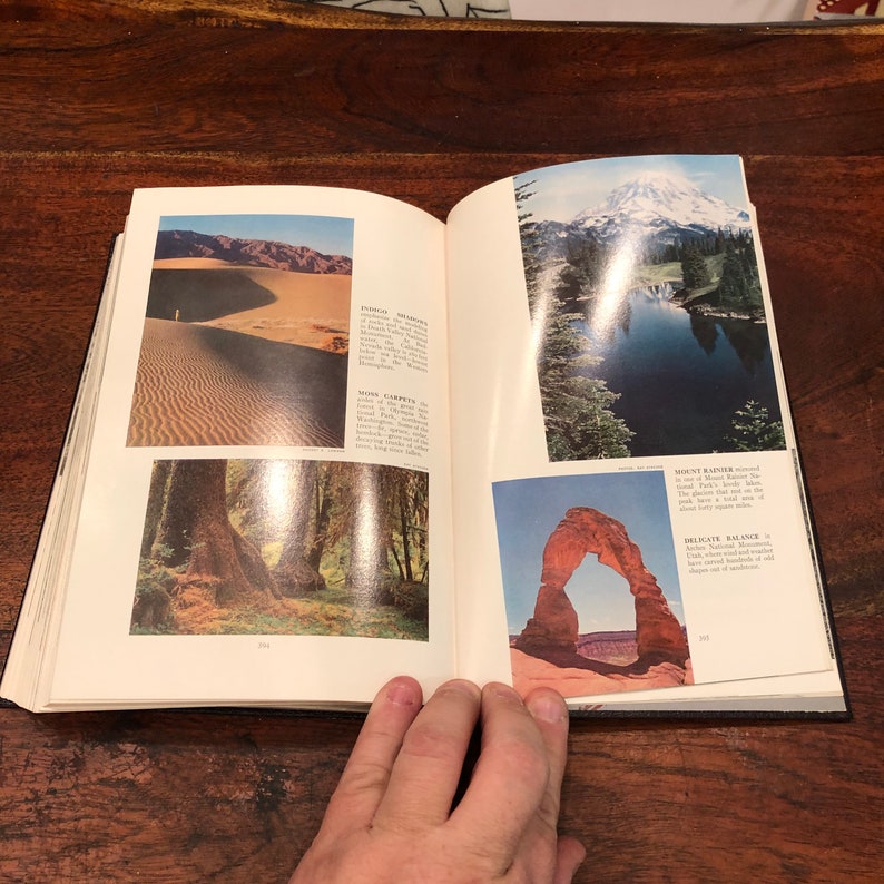 May include: An open book displaying colourful images of various landscapes. The book features pictures of sand dunes, a forest, a mountain with a lake, and a natural arch. The book's pages contain text describing the locations.