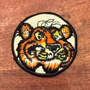 Op de afbeelding: Een ronde geborduurde patch met een gestileerd tijgergezicht in oranje, zwart en wit garen op een crèmekleurige stoffen achtergrond. Het ontwerp is omrand door een dikke, zwart gestikte rand en bevat gedetailleerde tijgerkenmerken.