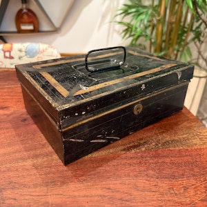 May include: A vintage black metal cash box with gold trim and a handle. The box has a keyhole and shows signs of wear, suggesting it is an antique. The cash box is rectangular and sits on a wooden surface.