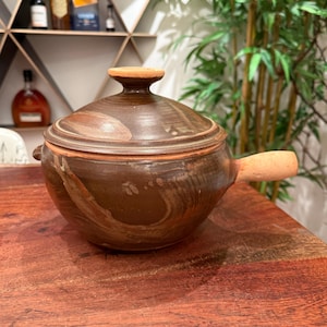 Puede incluir: Una cacerola de cerámica marrón con tapa y asa de madera. La cacerola tiene un aspecto rústico, hecha a mano, con un esmalte marrón y beige.