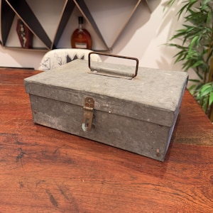 May include: A vintage metal box with a hinged lid and a metal handle. The box is painted a light grey colour and has a worn, rustic look. The box has a metal latch and a metal hinge.