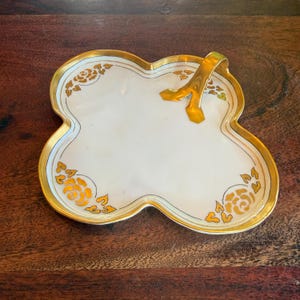 May include: A white, clover-shaped serving dish with gold trim and a gold handle. The dish features gold floral designs on the edges. The dish is likely made of porcelain or ceramic and is set on a dark wooden surface.