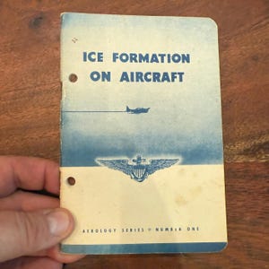Op de afbeelding: Vintage boekje getiteld "ICE FORMATION ON AIRCRAFT" met een blauw-witte omslag. De omslag toont een illustratie van een vliegtuig en een embleem. Het boekje wordt door een hand vastgehouden en heeft twee gaten om te binden. De tekst "AEROLOGY SERIES NUMBER ONE" staat onderaan gedrukt.