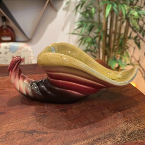 以下が含まれることがあります： 滑らかで光沢のある仕上げの陶器製のコーヌコピア型容器。この作品は、濃い茶色のベースから始まり、深い赤、明るいピンク、そして最後に淡い黄色の縁へと変化するグラデーションカラーが特徴です。