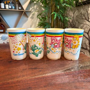 May include: Four vintage Care Bears plastic tumblers. Each tumbler features a different Care Bear character with rainbow and cloud designs. The tumblers have a white base with a rainbow stripe at the top and the Care Bears logo.