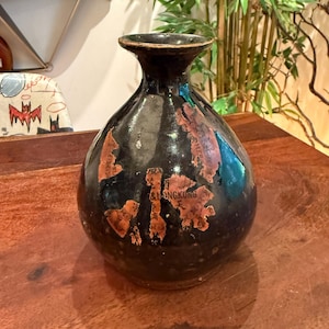 May include: A dark, antique vase with a rounded body and a slender neck. The vase is black with faded orange and brown labels, including the text "& HONGKUNG". The vase is likely ceramic and has a glossy finish.