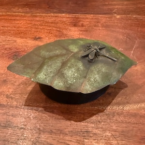 May include: A decorative metal leaf-shaped dish with a dragonfly on top. The leaf is green and gold with a textured surface. The dish sits on a black base. The dragonfly is silver-colored.