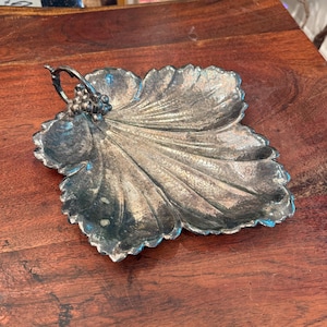 Puede incluir: Bandeja plateada en forma de hoja con asa. La bandeja tiene un diseño detallado de venas de hoja y un pequeño racimo de uvas en la base del asa. Se encuentra sobre una superficie de madera.