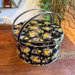 May include: A round, black tin with a floral pattern of yellow, purple, and white pansies. The tin has a silver handle and rim. The flowers are set against a black background. The tin is sitting on a wooden surface.