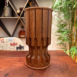 May include: A cylindrical wooden utensil holder featuring a dark brown finish and a series of vertical, recessed grooves carved into the exterior surface, creating a textured, ribbed appearance around the entire circumference of the object.