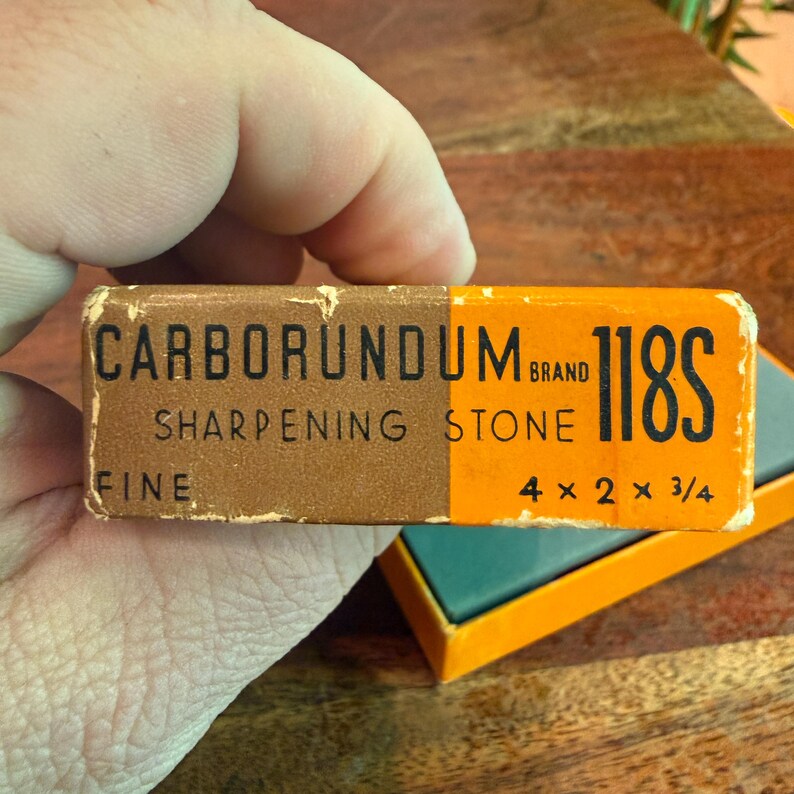 May include: A vintage Carborundum Brand 118S sharpening stone in its original box. The box is brown and orange, with the text "Carborundum Brand Sharpening Stone Fine" and dimensions of 4 x 2 x 3/4 inches.