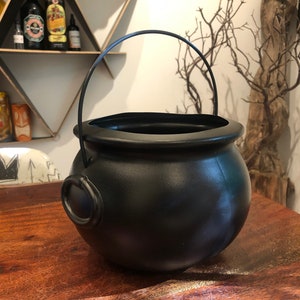 May include: A black cauldron with a curved handle and two side rings. The cauldron is on a wooden table. The cauldron is a decorative item.