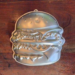 May include: A silver-colored, burger-shaped cake pan. The pan is made of metal and features detailed impressions of a hamburger, including the bun, patty, cheese, and lettuce. It has a small loop at the top for hanging.