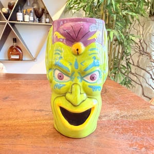 May include: A ceramic tiki mug with a green face, purple head, and yellow accents. The mug features a wide, open mouth, blue eyes, and a yellow nose. The mug is designed to resemble a monster or creature.