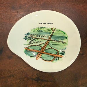 May include: An off-white, irregularly shaped ceramic dish featuring a hand-painted watercolor illustration of a highway interchange. The text "NEW YORK THRUWAY" is printed in black capital letters above the green and brown landscape graphic.