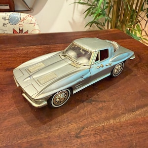 May include: A silver-coloured model car featuring a sleek coupe body style, detailed wheels, and a Fuel Injection emblem on the side. The vehicle has a split rear window design, a long bonnet with vents, and clear windows revealing a dark interior cabin.