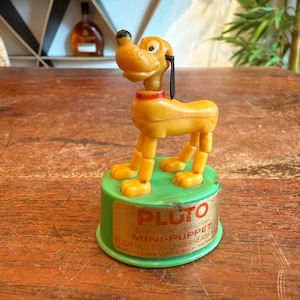 Op de afbeelding: Een vintage Pluto mini-poppetje. De plastic speelgoedhond is geel met een zwart oor, een rode kraag en een zwarte neus. Het speelgoed staat op een groene basis met de naam "PLUTO" erop gedrukt. De tekst "MINI-PUPPET" en "PUSH BUTTON UNDERNEATH" is ook zichtbaar.