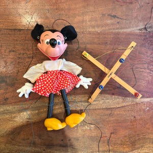 Puede incluir: Una marioneta vintage de Minnie Mouse con una falda de lunares rojos y blancos, guantes blancos y zapatos amarillos. La muñeca tiene orejas negras y una cruz de control de madera. La muñeca está sobre una superficie de madera.