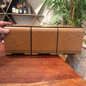 May include: A rectangular wooden storage unit featuring three front-facing drawers with a light oak wood grain finish. Each drawer is accented with two thin horizontal black lines near the base and a black trim along the top edge.