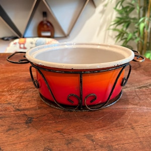 Pode incluir: Uma pequena panela redonda esmaltada com um gradiente vibrante de laranja e vermelho. A panela tem um interior branco e é sustentada por um suporte de ferro forjado preto com redemoinhos decorativos. A panela tem duas alças.