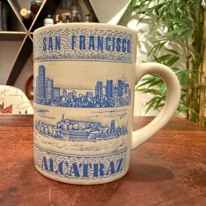 May include: A cream-colored ceramic mug featuring blue line-art illustrations. The top section displays the text SAN FRANCISCO above a city skyline. The lower section shows an island landscape with the text ALCATRAZ printed at the bottom.