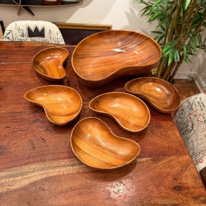 Puede incluir: Conjunto de seis cuencos de madera de diferentes tamaños. Los cuencos son todos de forma de riñón y tienen un acabado de madera natural. Los cuencos están dispuestos sobre una mesa de madera.