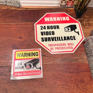 May include: Two warning signs on a wooden surface. The larger sign is red with white lettering and says "WARNING 24 HOUR VIDEO SURVEILLANCE TRESPASSERS WILL BE PROSECUTED". The smaller sign is silver with yellow lettering and says "WARNING 24 HOUR Video Surveillance".