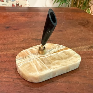 May include: A desk pen holder featuring a polished, cream-colored onyx stone base with natural tan veining. A brass-toned metal swivel joint connects the base to a tapered, black cylindrical pen socket angled upward for holding a writing instrument.
