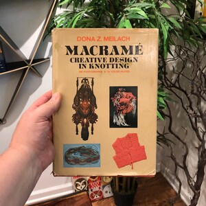 May include: A book titled "Macrame Creative Design in Knotting" by Dona Z. Meilach. The book cover features three macrame projects: a hanging wall decor, a headpiece, and a vest. The book cover is yellow with brown text and has 368 photographs and 18 colour plates.