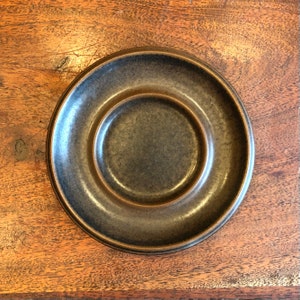 May include: A dark brown ceramic saucer with a slightly raised center. The saucer has a smooth, glossy finish.