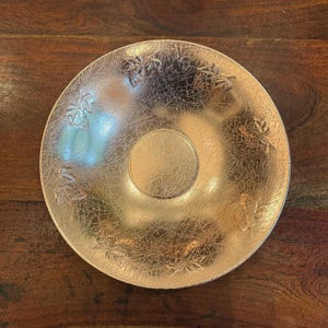 May include: A round, gold-colored decorative plate with a textured surface and a central, smaller circular indentation. The plate features leaf-like designs around the edge.