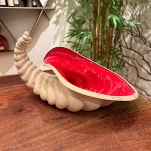 May include: A ceramic decorative bowl in the shape of a conch shell. The exterior is a cream color, and the interior is a glossy red. The bowl is placed on a wooden surface.