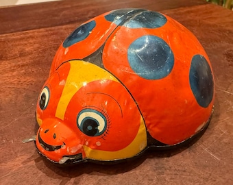 May include: A vintage tin toy shaped like a ladybug with a bright orange body, yellow head accents, and large blue circular spots. The toy features a cartoonish face with wide eyes, long eyelashes, and a smiling mouth, with small metal antennae.