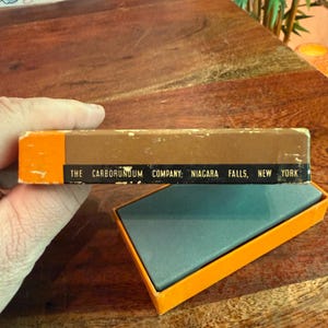 May include: A vintage Carborundum Company sharpening stone in its original box. The box is brown and orange with the text "THE CARBORUNDUM COMPANY: NIAGARA FALLS, NEW YORK". The open box reveals a gray stone.