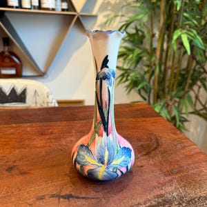 May include: A small, white ceramic vase with a scalloped rim. The vase is decorated with a pink, blue, and green floral design featuring irises.
