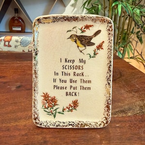 May include: Rectangular ceramic scissor holder with a bird and floral design. The cream-colored holder has gold trim and text that reads, "I Keep My SCISSORS In This Rack... If You Use Them Please Put Them BACK!"