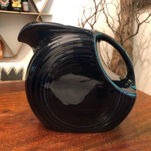 May include: A navy blue ceramic pitcher with a circular handle and a spout. The pitcher has a glossy finish and a ridged design around the body. It sits on a wooden surface.