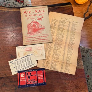 Op de afbeelding: Vintage ephemera met een Air-Rail Stationery boekje met een treinillustratie, een lijst, enveloppen en een War Bonds advertentie. Er is ook een kleine rode en blauwe postkaart aanwezig.