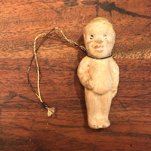 May include: A vintage porcelain doll ornament with a white body, a yellow head, and black painted eyes. The doll is wearing a black necklace and has a string attached to its head.