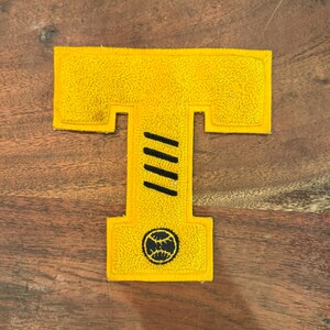 May include: A yellow felt letter "T" patch with a textured surface. The letter features three black diagonal stripes and a baseball graphic. The patch is likely for a varsity jacket or similar apparel.