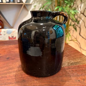 May include: A dark, glossy black ceramic jug with a rounded body, a short neck, and a single curved handle attached to the side. The surface of the jug is smooth and reflective, showing light highlights across its curved, cylindrical form.