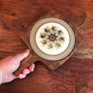 May include: A wooden serving board with a round ceramic plate featuring a floral design in shades of green, yellow, and brown. The plate has a white background and a raised center with a brown circle.