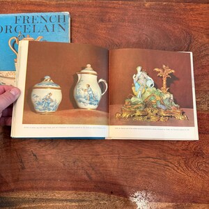 May include: Open book with images of antique French porcelain. The left page shows a small jug and sugar bowl with blue and white landscape scenes. The right page displays a decorative statue with gold accents.