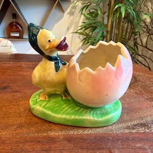 May include: A ceramic duck figurine wearing a bonnet and a bow, next to a cracked eggshell planter. The duck is yellow with a dark green bonnet and bow, and the eggshell is pink and white. The base is green.