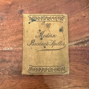 May include: A vintage book with a tan cover and black text that reads "Modern Business Speller". The cover has a decorative border of leaves and is stained with dark brown spots.