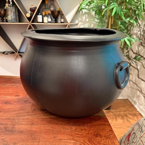 Peut inclure: Un grand chaudron noir avec un corps arrondi et un large bord. Il comporte une poignée sur un côté. Le chaudron est fait d'un matériau mat et repose sur une surface en bois. L'arrière-plan comprend une étagère avec des bouteilles et des plantes décoratives.